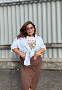 Witte geknoopte blouse over een graphic tee, gecombineerd met een bruine wikkelrok. Accessoires omvatten een zonnebril en ringen, met een gladde textuur.