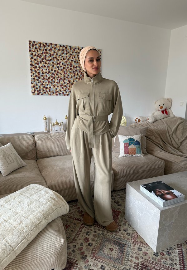 Mujer con mono beige y pañuelo en la cabeza, de pie en un salón con sofá beige, alfombra con diseño y cojines decorativos.