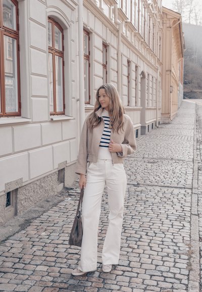 Joven mujer con chaqueta beige y pantalones blancos de pie en una calle adoquinada junto a un edificio color crema con grandes ventanas.