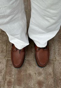 Brune læder bådsko med en tekstureret finish, sorte gummisåler og sorte snørebånd, kombineret med hvide linnedbukser. I en murstensomgivelse.