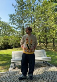 Πλεκτό πουλόβερ σε μπεζ χρώμα με γραφικό φλόγας και κίτρινη λεπτομέρεια. Συνδυασμένο με φαρδιά τζιν παντελόνια σε σκούρο χρώμα και μαύρα παπούτσια. Φυσικό φόντο.