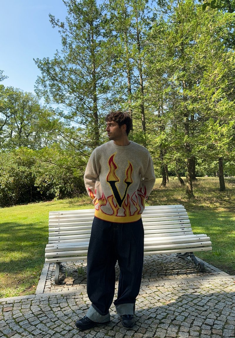 Πλεκτό πουλόβερ σε μπεζ χρώμα με γραφικό φλόγας και κίτρινη λεπτομέρεια. Συνδυασμένο με φαρδιά τζιν παντελόνια σε σκούρο χρώμα και μαύρα παπούτσια. Φυσικό φόντο.