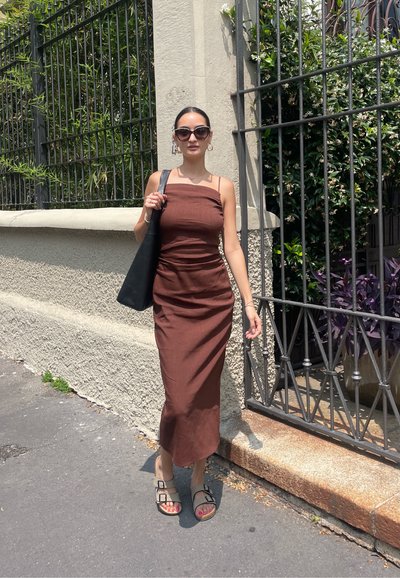 Vestido midi marrón, ajustado, con tirantes finos, de tejido texturizado, combinado con un tote negro y sandalias con dos hebillas. Vegetación al fondo.