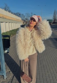 Jachetă crem din blană artificială cu mâneci voluminoase, hanorac bej dedesubt, șapcă roz deschis și geantă maro țesută, pe fundal urban.