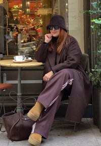 Manteau marron, pantalon large assorti avec des rayures blanches, bottes beige et chapeau en feutre. Assis à une table de café avec une tasse et une pâtisserie.