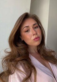 Joven mujer con el pelo largo y ondulado de color castaño, lleva lápiz labial rosa y una camiseta rosa pálido, posando en interiores frente a una pared bicolor.