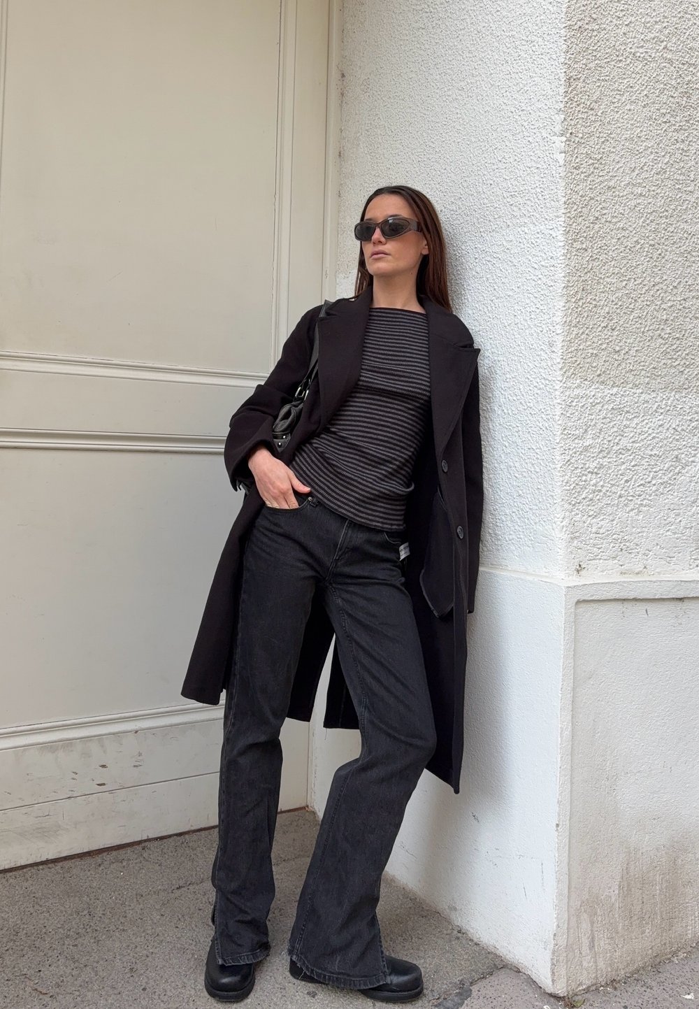 A black wool coat worn over a black and white striped top, teamed with dark flared jeans and chunky black boots. Accessories feature black sunglasses.