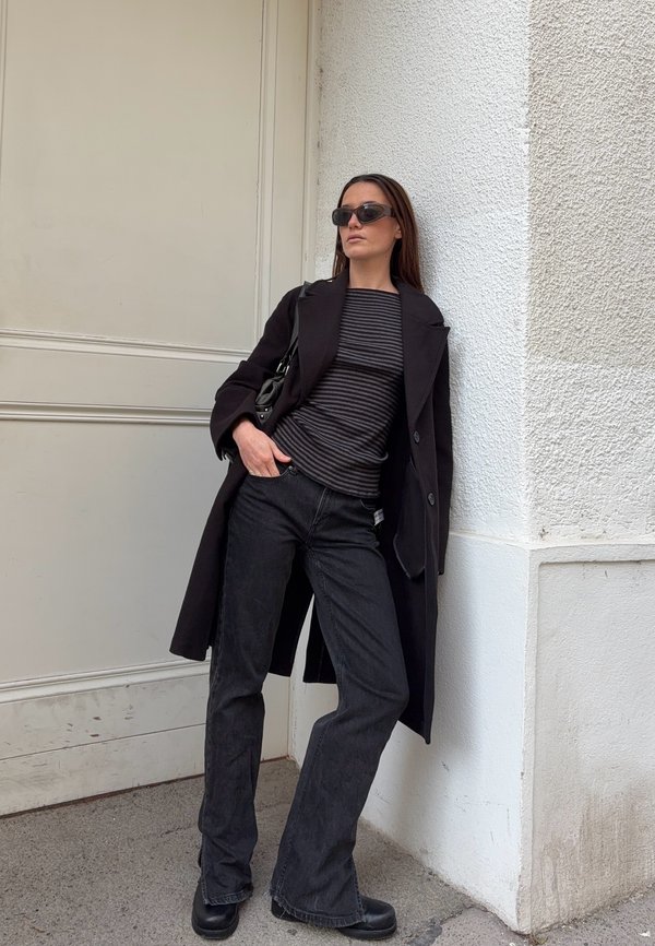 Cappotto di lana nero sopra una maglietta a righe bianca e nera, abbinato a jeans svasati scuri e stivali neri spessi. Gli accessori includono occhiali da sole neri.