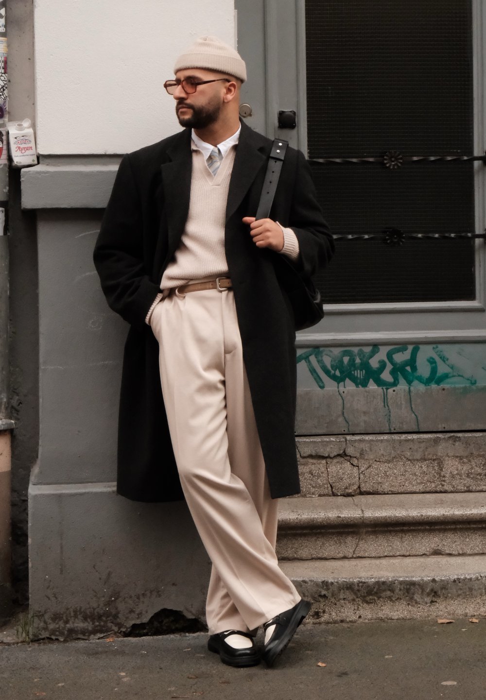 Μαύρο παλτό, μπεζ πλεκτό πουλόβερ, μπεζ παντελόνι, μαύρη ζώνη, στρογγυλά γυαλιά και ανοιχτό μπεζ σκουφί. Μαύρα παπούτσια με λευκές λεπτομέρειες.