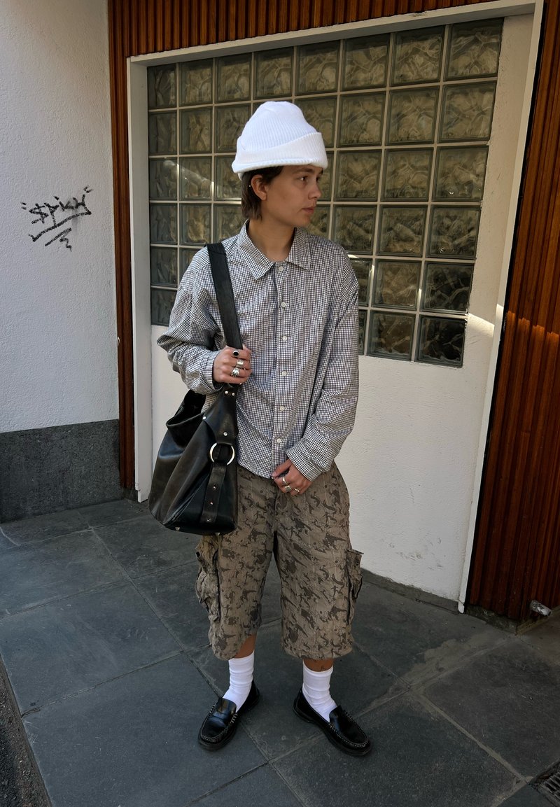 Personne portant un bonnet blanc, une chemise à carreaux, un short cargo à motifs, des chaussettes blanches, des chaussures noires, portant un grand sac à bandoulière noir, debout sur le trottoir.