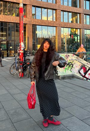 Chaqueta bomber de cuero negro con cuello de piel, falda de lunares, bolso rojo y zapatos rojos. De pie afuera cerca de bicicletas y un edificio moderno.