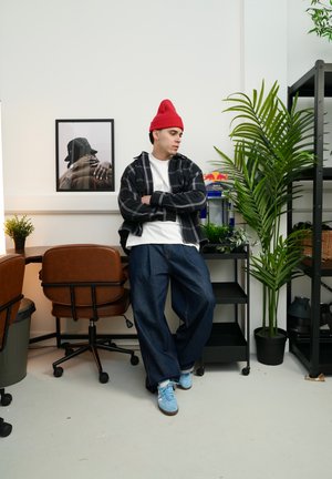 Joven con gorro rojo, camisa de cuadros y vaqueros anchos apoyado en un carrito negro en una habitación moderna con plantas y sillas de oficina marrones.