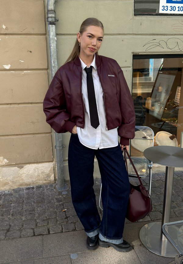 Tânăra stă pe stradă purtând o jachetă bomber burgund, cămașă albă cu cravată neagră, blugi largi și închiși la culoare, pantofi negri, ținând o geantă burgund.