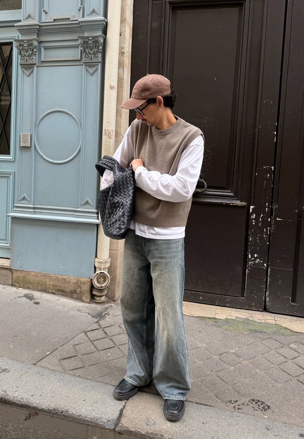 Man adjusting black woven bag, wearing brown cap, glasses, beige sleeveless jumper over white long-sleeve shirt, loose jeans, standing by a blue door.