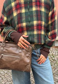 Personne portant une veste à carreaux verte, rouge et jaune avec des manches marron, un jean bleu clair, tenant un sac à main en cuir marron, montrant des bagues aux doigts.