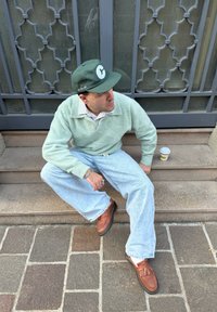 Suéter verde menta sobre uma camisa às riscas, jeans baggy azuis claros, mocassins castanhos, segurando um copo de café, sentado nas escadas de pedra ao lado de um portão decorativo.