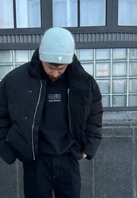 Doudoune noire avec fermeture éclair, portée sur un sweat-shirt noir avec un motif blanc. Bonnet tricoté bleu clair à texture.