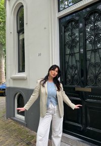Light beige blazer worn over a blue button-up shirt and white patterned trousers. A black ornate door featuring decorative ironwork and the house number 10.