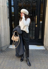 Γυναίκα με κρεμ πουλόβερ, λευκό μπέρε, μαύρο παντελόνι και μπότες ρυθμίζει τα γυαλιά ηλίου, κρατώντας σκοτεινό παλτό και μπεζ τσάντα δίπλα σε μια μαύρη σιδερένια πόρτα.