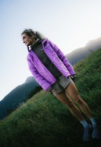 Jacke in helllila mit Steppung und hohem Kragen, über einem dunkelbraunen Oberteil und beigen Shorts getragen, vor einer grünen Landschaft und Bergen.