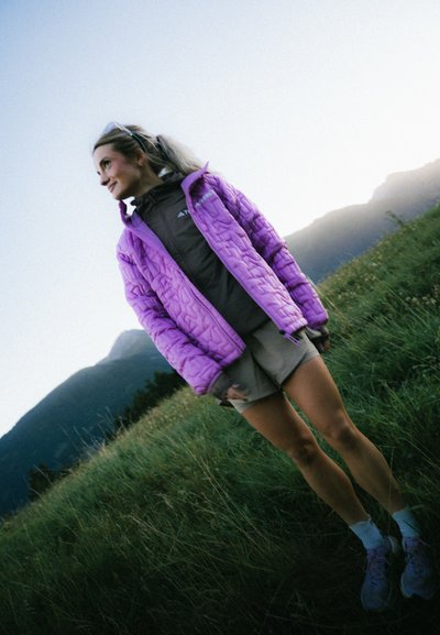 Chaqueta acolchada de color púrpura claro con cuello alto, superpuesta sobre una blusa marrón oscuro y pantalones cortos beige, situada frente a un paisaje de hierba y montañas.