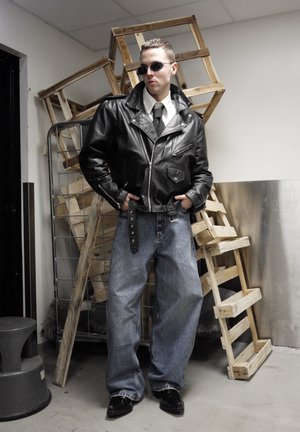 Chaqueta de cuero negra para motocicleta con cremalleras, camisa blanca, corbata negra y vaqueros azules holgados. Zapatos negros, gafas de sol; al fondo, pilas de madera.