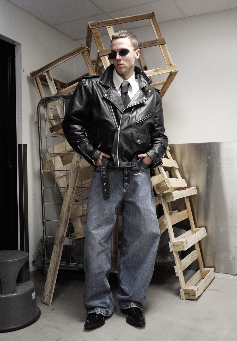 Veste de moto en cuir noir avec des fermetures éclair, chemise blanche, cravate noire et jean bleu ample. Chaussures noires, lunettes de soleil ; en arrière-plan, des piles de bois.