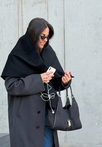 Crni kaput s velikom, prebačenom crnom šalom. Drži bijeli pametni telefon s slušalicama, crnu torbu sa srebrnim detaljima i traperice.