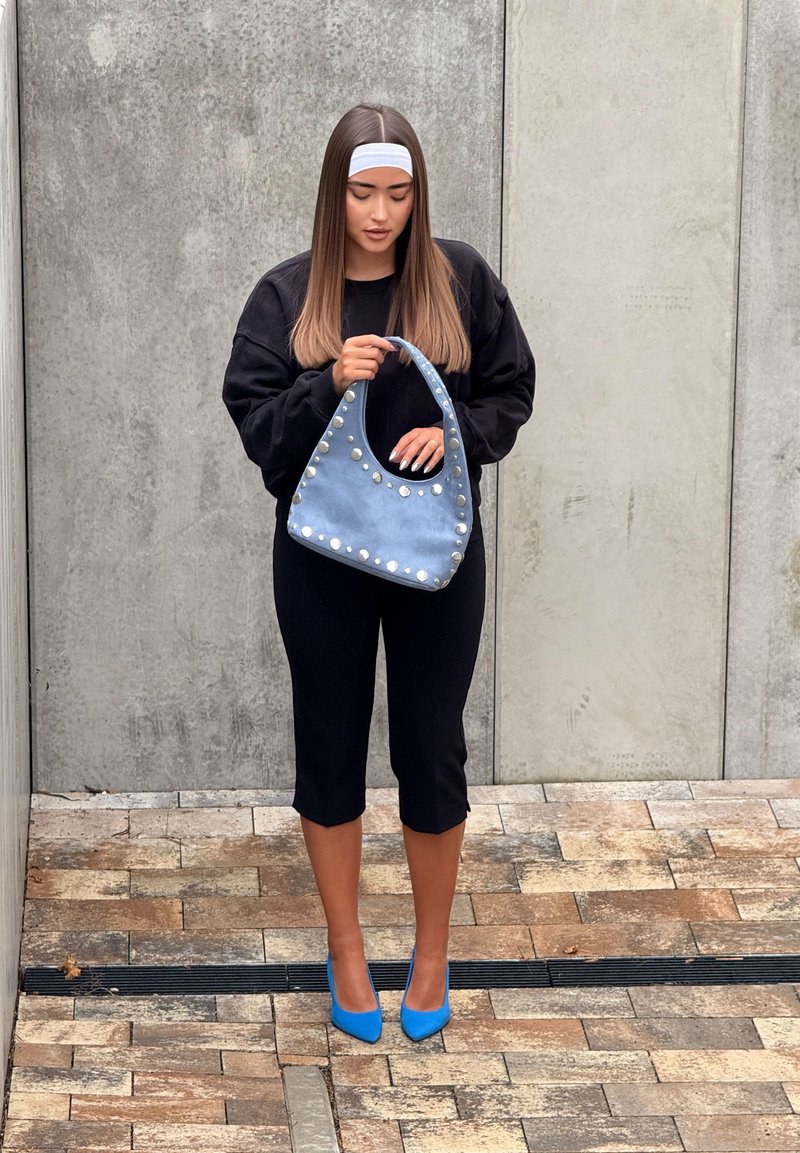 Sac à main en daim bleu orné de clous argentés, porté avec un pull noir oversize et un pantalon noir court, complété par des escarpins bleus.