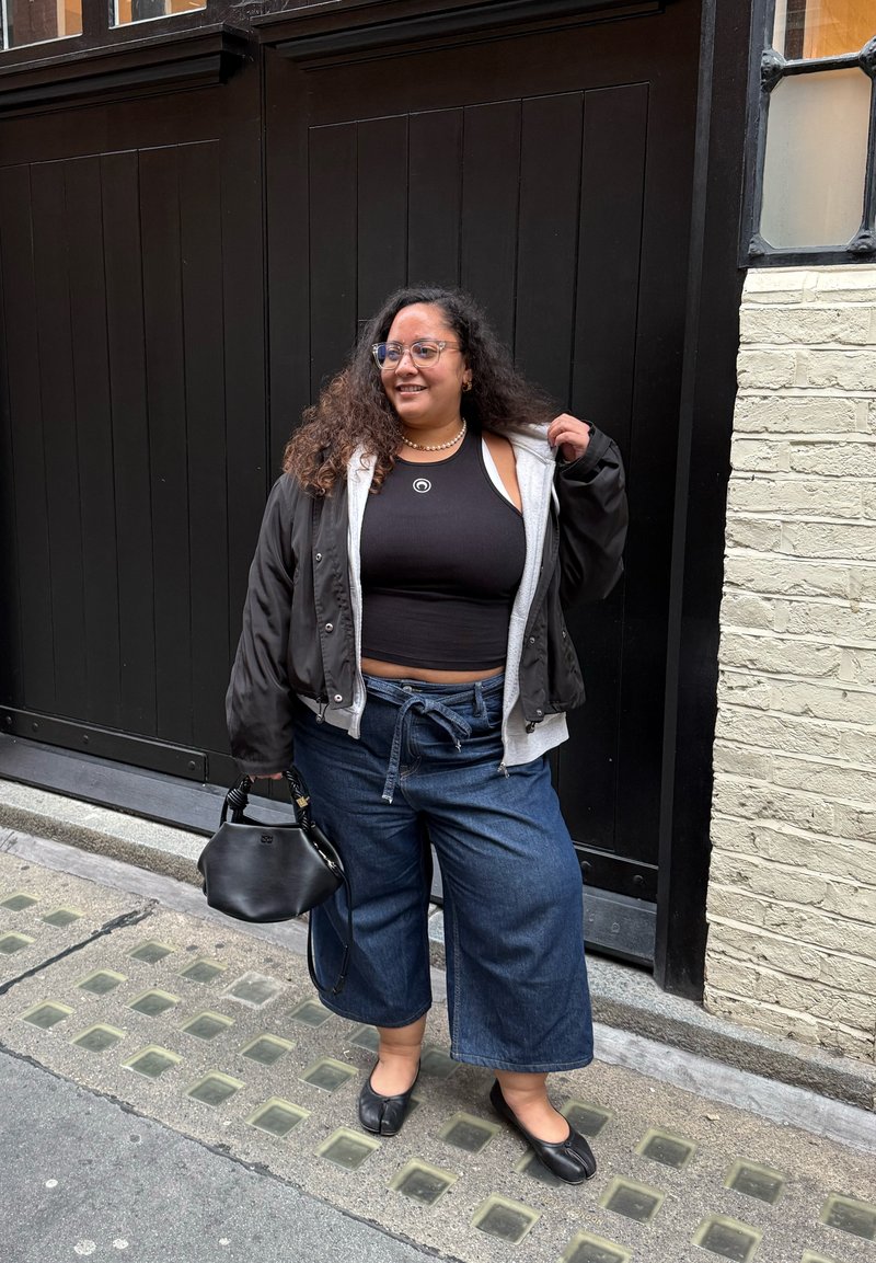 Black bomber jacket over a black tank top, wide blue denim pants, black flats, and a small black handbag with a textured handle.