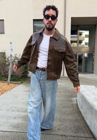 Veste en cuir marron avec poches poitrine, T-shirt blanc et jean bleu clair ample. Le mannequin porte des lunettes de soleil et se tient sur un chemin en béton.