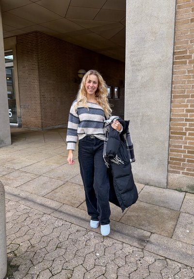 Jersey de punto a rayas en tonos gris y azul marino, vaqueros anchos negros, zapatos azul claro, sosteniendo un abrigo acolchado negro, de pie sobre un pavimento de ladrillo.