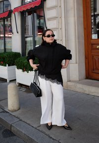 Mustapohjainen ylisuuri paita pitsiyksityiskohtineen, valkoiset leveälahkeiset housut, mustat kärkitossut ja pieni musta laukku, kaupunkimaisemaa vasten.
