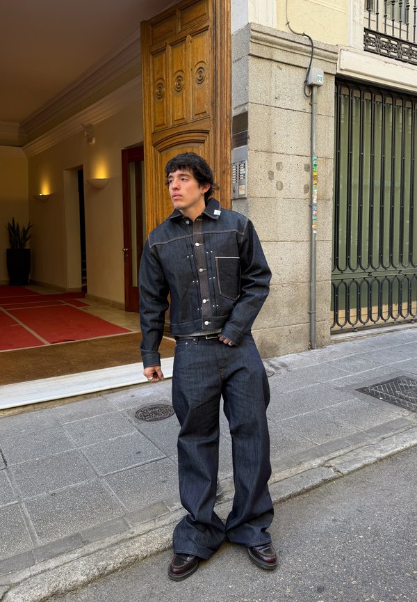 Ung mand iført mørk denimjakke og brede jeans står på en byfortov nær en bygning med en trædør.