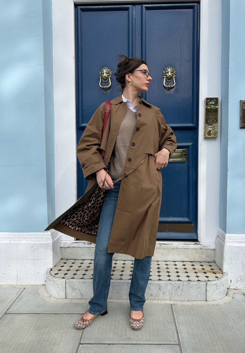 Abrigo marrón de trench con un interior forrado de leopardo, suéter beige, jeans azules y bailarinas con estampado. De pie junto a una puerta azul con un aldaba de león.