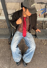 Osoba u crnoj bomber jakni preko smeđe košulje, crvene kravate i širokih svijetloplavih traperica, sjedi na sivom klupcu. Crne cipele i kapa.