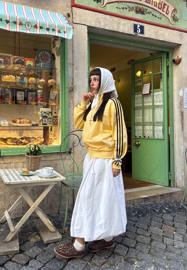 Giovane donna con giacca gialla e gonna bianca in piedi fuori da una panetteria con porta verde, con una vetrina di pane e dolci, accanto a un tavolo con tè.