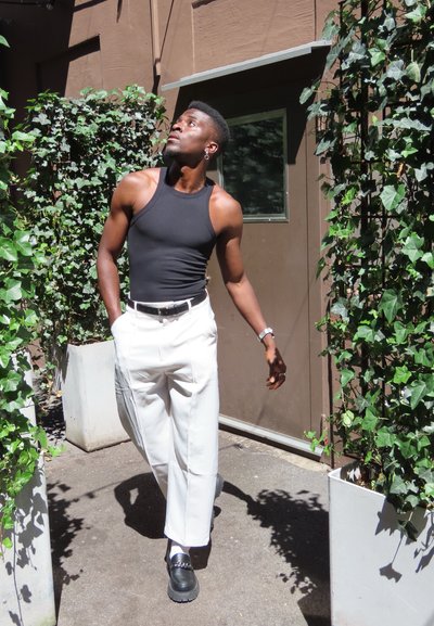 Top negro sin mangas, pantalones blancos de pierna ancha y zapatos negros con suelas gruesas. Rodeado de hiedra verde y una pared marrón. Luz diurna.