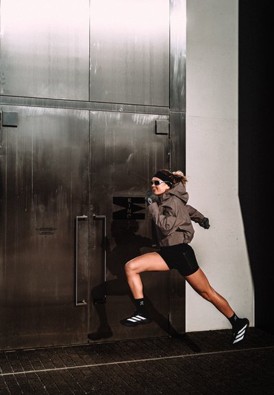 Mujer con gafas de sol y ropa deportiva corre en pleno paso junto a grandes puertas metálicas que proyectan una sombra nítida en la pared.