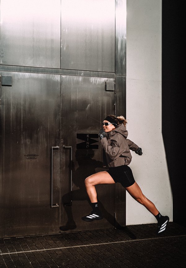 Napszemüveget és sportos ruházatot viselő nő fut középtempóban egy nagy fém ajtók előtt, amelyek éles árnyékot vetnek a falra.