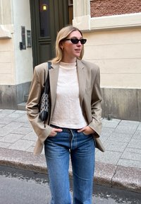 Ανοιχτό μπεζ υπερμεγέθη σακάκι πάνω από μια υφασμάτινη κρέμα μπλούζα, συνδυασμένο με ανοιχτό μπλε καμπάνα παντελόνι. Μαύρη τσάντα ώμου με λογότυπο και σκούρα γυαλιά ηλίου.