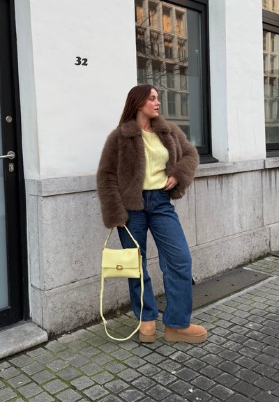 Chaquetón de piel sintética marrón, suéter de punto amarillo, jeans azules, bolso amarillo y botas de plataforma beige, de pie contra una pared y ventana blancas.