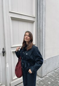 Zestaw skórzany z dżinsową kurtką i spodniami w ciemnym niebieskim kolorze, z pionowym żebrowanym wzorem. Posiada pasek w talii, duże przednie kieszenie i guziki. Czerwona torebka.