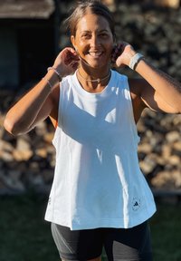 Femeie zâmbitoare își ajustează cerceii în aer liber, purtând un top alb fără mâneci, pantaloni scurți negri, un ceas de fitness și brățări în lumina soarelui.