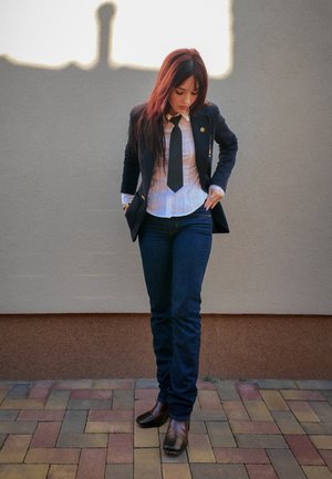 Blazer negro con botones dorados, camisa blanca de rayas, corbata negra, jeans azul oscuro y botas de tobillo marrones; de pie contra una pared texturizada.