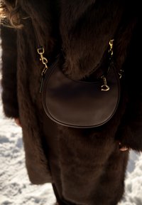 Άτομο που φοράει σκούρο καφέ παλτό από γούνα, κρατώντας μια σκούρα καφέ δερμάτινη τσάντα με χρυσά μεταλλικά στοιχεία, στέκεται σε χιονισμένο έδαφος.