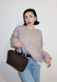Femme en pull beige duveteux et jeans bleu clair tenant un sac à main tissé brun foncé, posant devant un fond blanc uni.