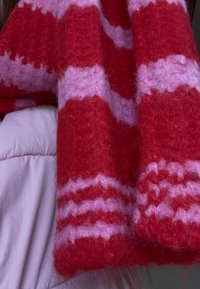 Neulottu huivi, jossa on vuorotellen punaisia ja vaaleanpunaisia raitoja, karhea tekstuuri. Mallissa on aaltoilevia ja tasaisia osioita. Pehmeä, lämmin materiaali.