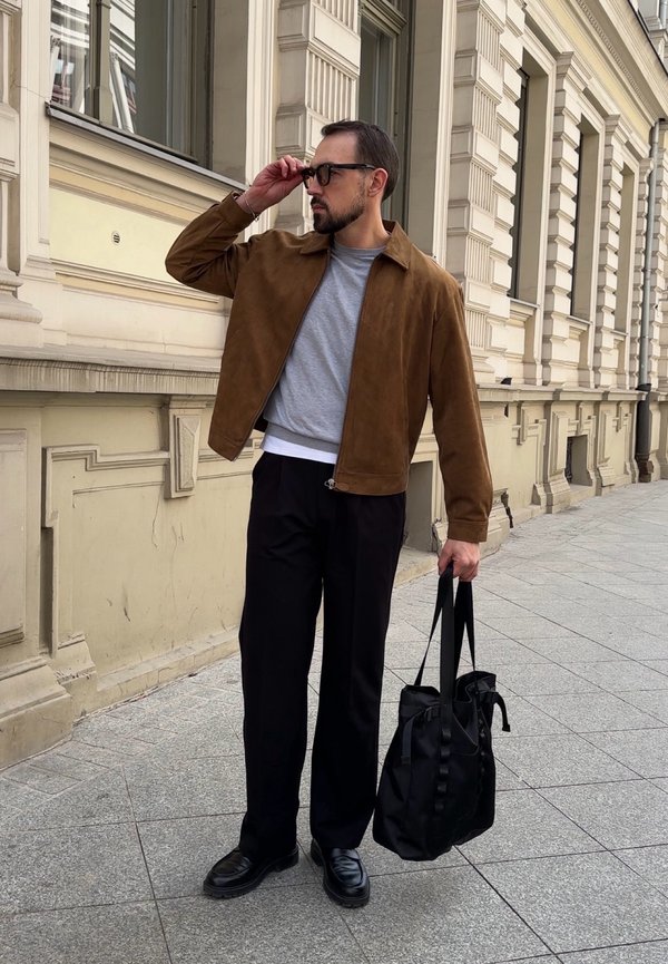 Homme en veste marron ajustant ses lunettes de soleil, tenant un sac fourre-tout noir, debout sur un trottoir urbain à côté d'un bâtiment beige orné.