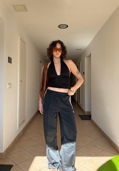 Top corto negro tipo halter, pantalones negros sueltos con rayas blancas, bolso marrón. Cabello rizado, gafas de sol naranjas, de pie en un pasillo.